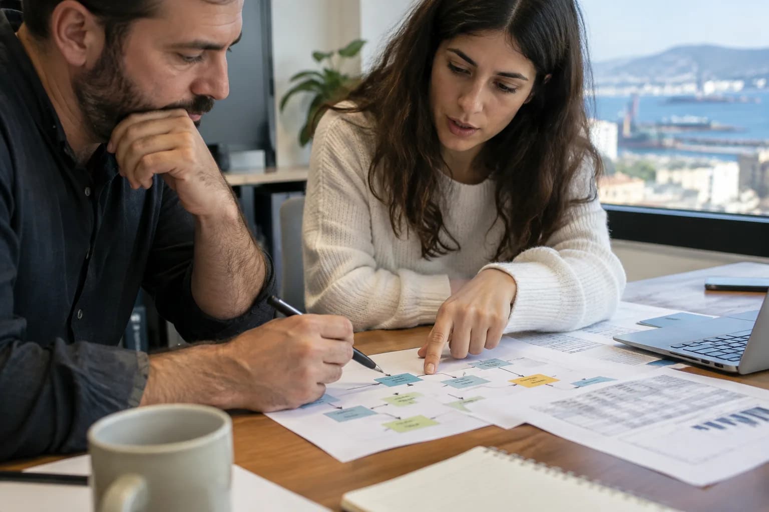 שני מייסדי סטארטאפ בחדר ישיבות קטן בחיפה, בוחנים תרשים תהליך עבודה ודפי הוצאות מודפסים