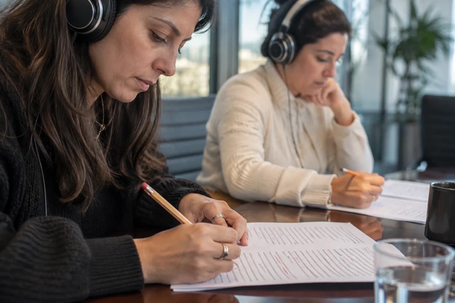 שתי בודקות עם אוזניות יושבות בחדר ישיבות מול דפי תסריט ומסמנות תיקוני הגייה בעיפרון