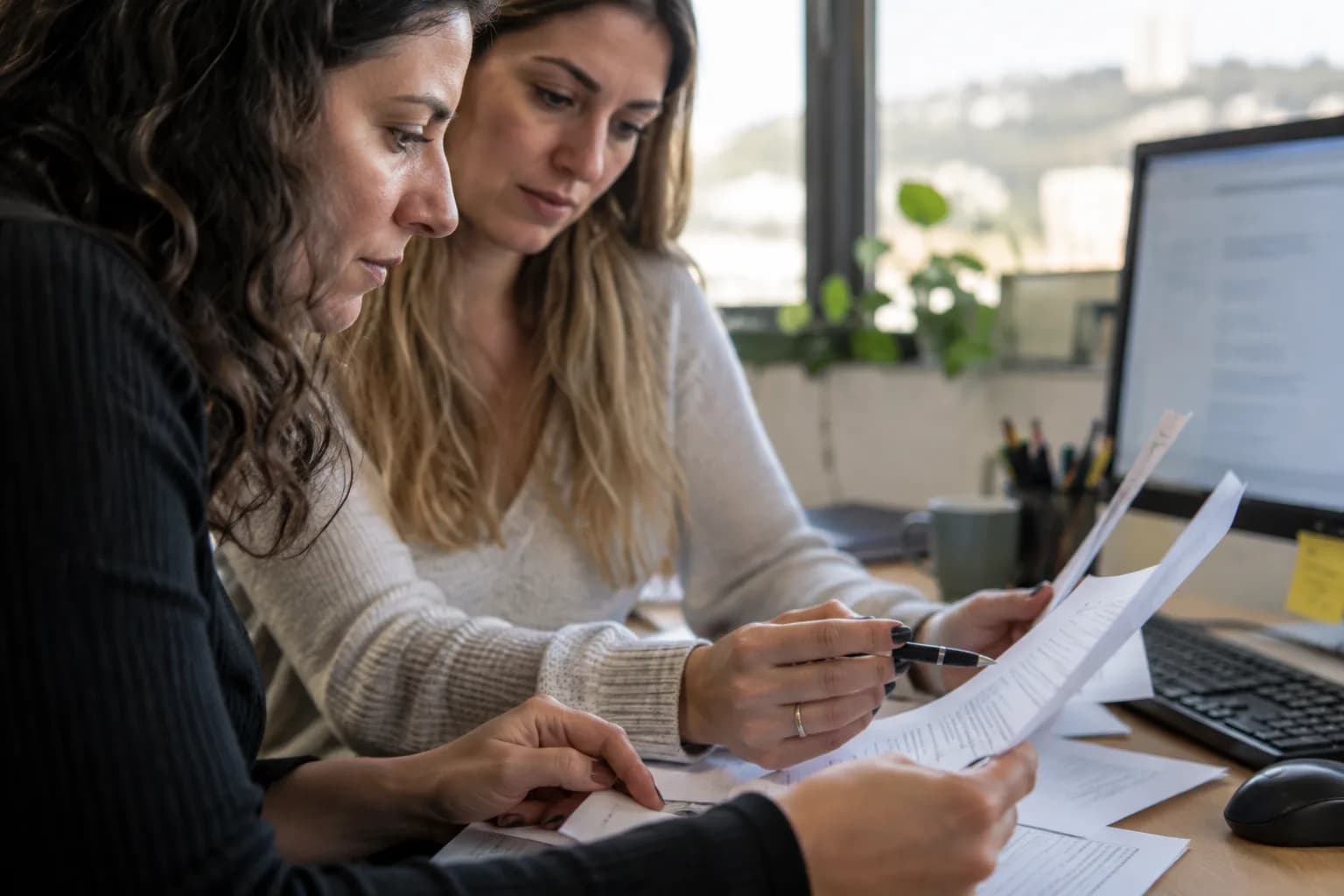 שתי עובדות במשרד קטן בחיפה בוחנות על דפים מודפסים גרסאות של תשובה ללקוח, לצד מסך מטושטש
