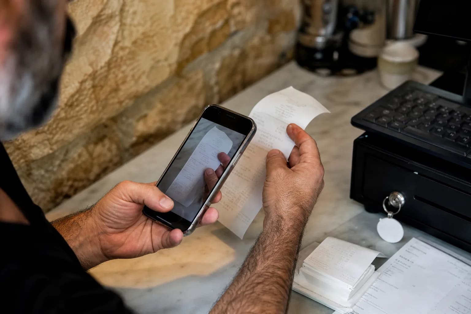 בעל מסעדה בירושלים סורק חשבוניות בסמארטפון ליד דלפק הקופה כחלק מתהליך דיגיטציה