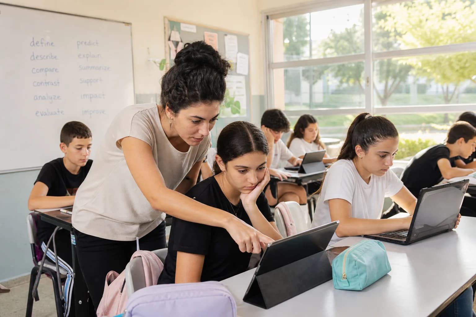 תלמידי חטיבת ביניים בישראל לומדים עם מחשבים ניידים בכיתה, מורה רוכנת לעזור לתלמיד